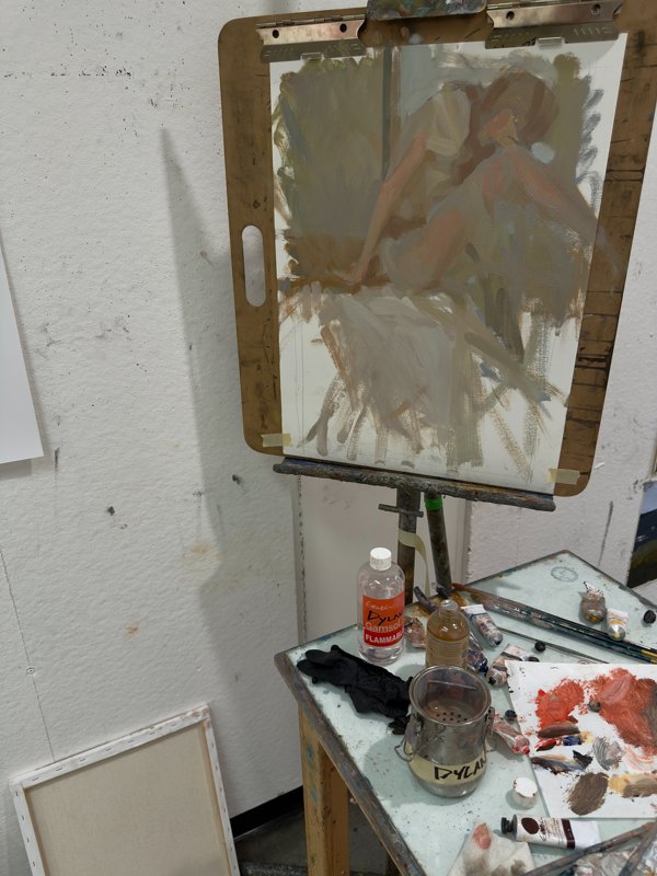 A painting in progress on an easel, surrounded by palette knives, paint tubes, and studio supplies